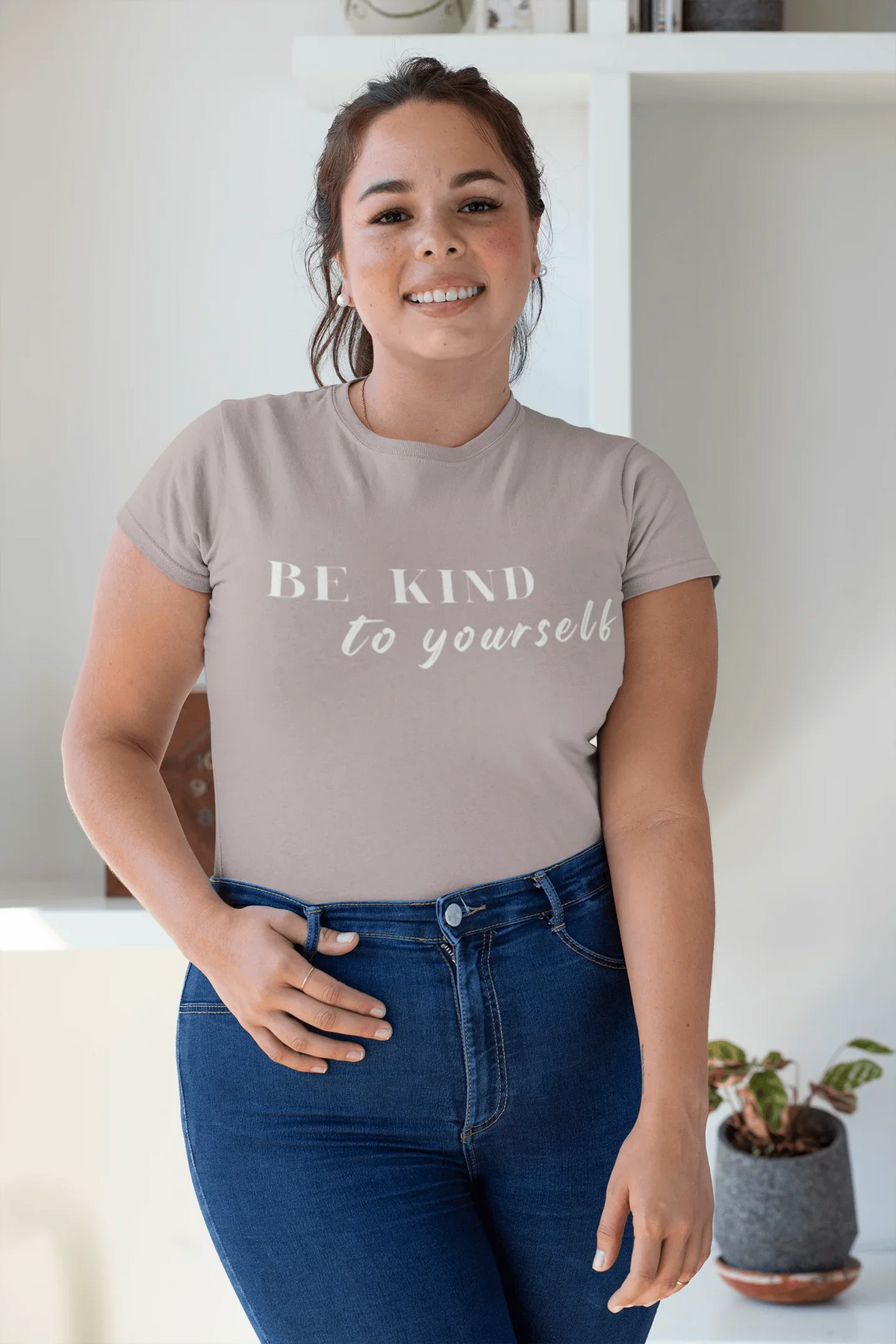 A woman wearing a red V-neck t-shirt with the text 'BE KIND to yourself' in white script, paired with light-wash denim shorts, set against a solid off-white background.