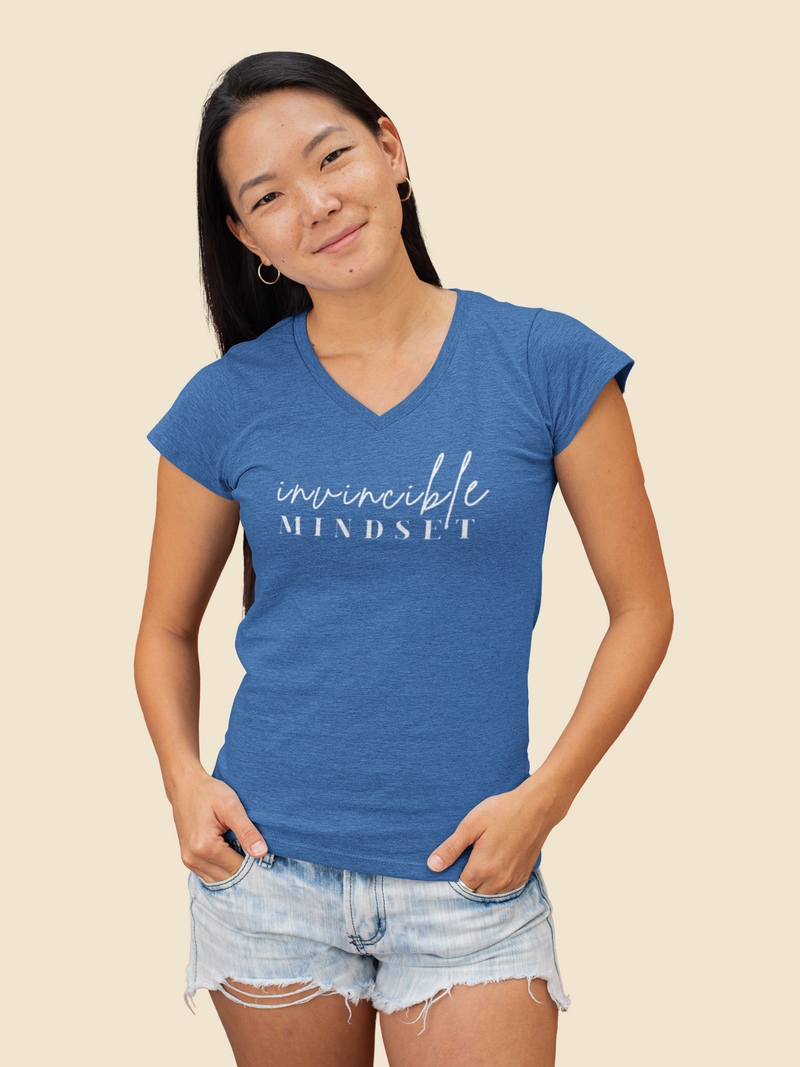 A woman modeling a heather blue V-neck t-shirt with 'invincible MINDSET' printed in white script across the chest, paired with light-wash denim shorts against a solid beige background.