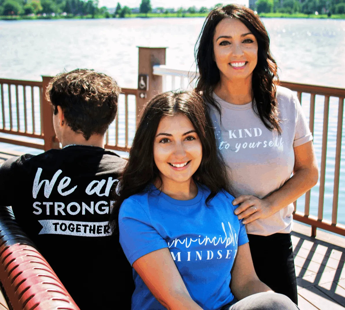 Founders Antonette, Brandon, and Lauryn, wearing graphic t-shirts with inspirational quotes are pictured outdoors on a wooden dock with water in the background. Lauryn in the front wears a bright blue t-shirt that reads 'invincibly MINDSET' in white script.