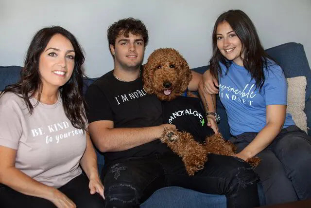 Founders of Always Love Being and a brown poodle sitting together on a blue couch, wearing matching graphic t-shirts with the text 'I'M NOT ALONE, Be kind to yourself, and Invincible Mindset