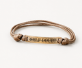 A braided tan rope bracelet with a central gold-toned metal bar engraved with the words 'SELF-DOUBT' in black capital letters, set against a plain white background.