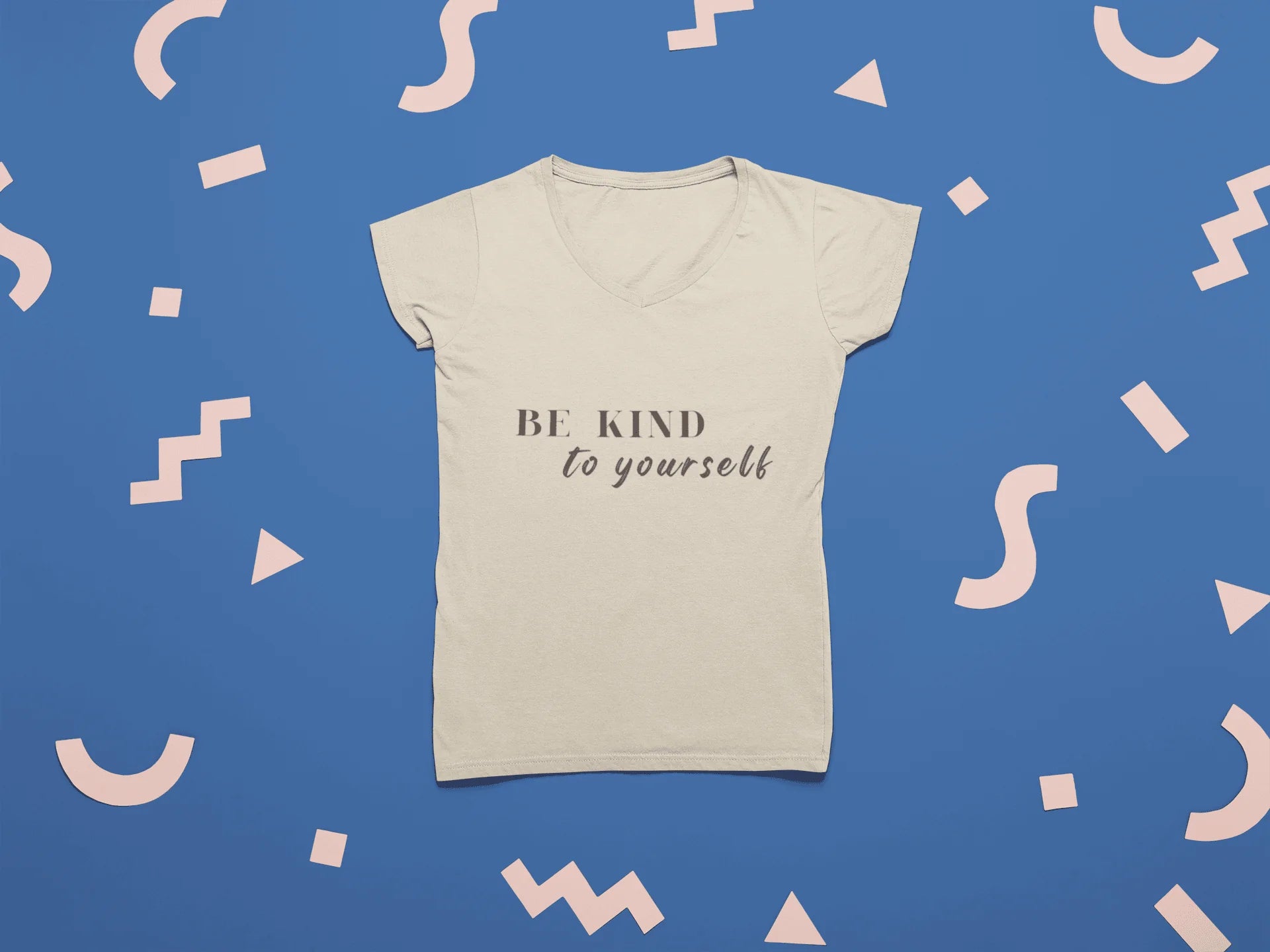 A cream-colored short-sleeve V-neck t-shirt with the text 'BE KIND to yourself' printed on the chest, displayed flat against a blue background decorated with pastel pink geometric shapes.