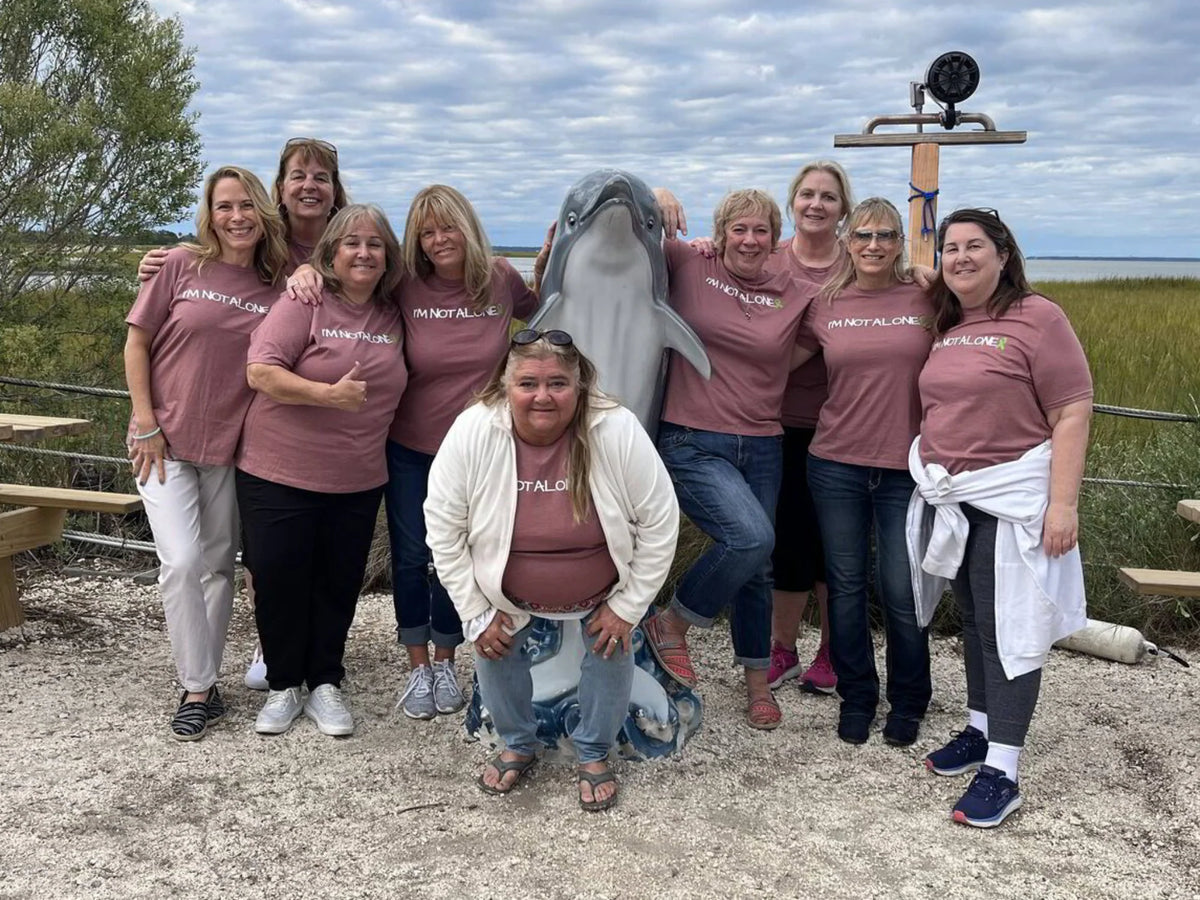 A group of nine women posing outdoors with a dolphin figurine in the center. They are wearing matching dusty rose t-shirts with white text that reads 'I AM NOT ALONE'.