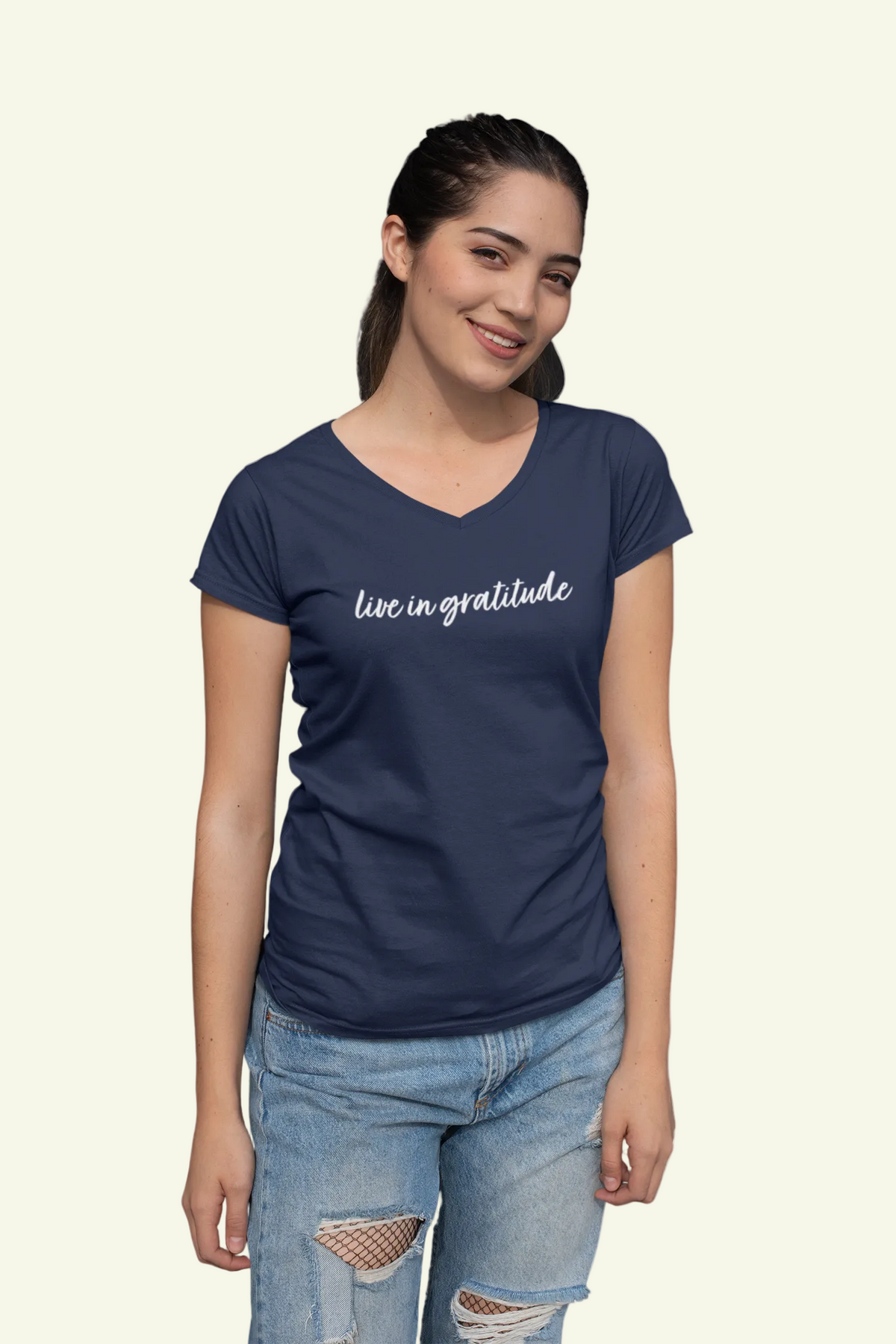 A woman wearing a navy blue v-neck t-shirt with 'live in gratitude' printed in white script across the chest, paired with light-wash distressed jeans, set against a plain light gray background.