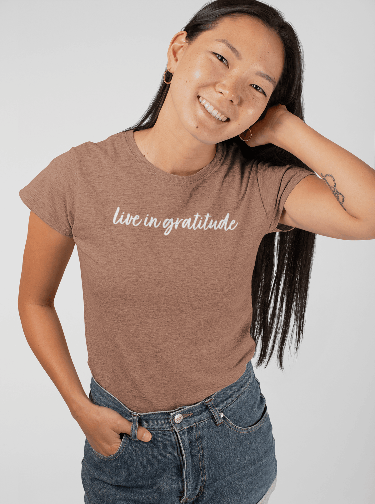A woman wearing a chestnut t-shirt with 'live in gratitude' printed in white script across the chest, paired with blue jeans, against a light gray studio background.