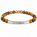 A beaded stretch bracelet with round brown and gold-toned stone beads, a small silver bead, and a polished silver bar engraved with the words 'Live in Gratitude'.