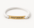 A white cord bracelet with a gold-plated bar engraved with the word 'SELF-DOUBT' in black capital letters, set against a plain white background.