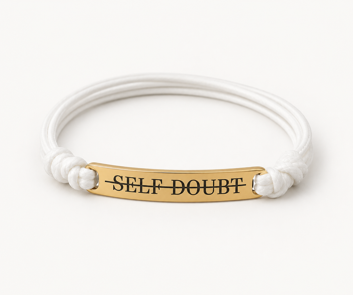 A white cord bracelet with a gold-plated bar engraved with the word 'SELF-DOUBT' in black capital letters, set against a plain white background.