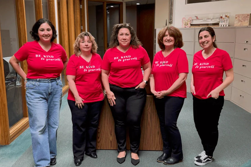 Five women standing in an office hallway wearing matching bright red t-shirts with white text that reads Be Kind To Yourself