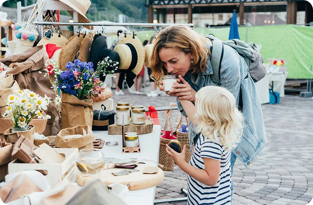 A woman in a light blue denim jacket and a young blonde girl in a navy and white striped shirt browse a rustic outdoor market stall filled with straw hats, bouquets, and small jars.