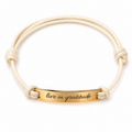 A double-strand cream-colored cord bracelet with a polished gold-plated ID bar engraved with the words 'live in gratitude' in black script, set against a plain white background.
