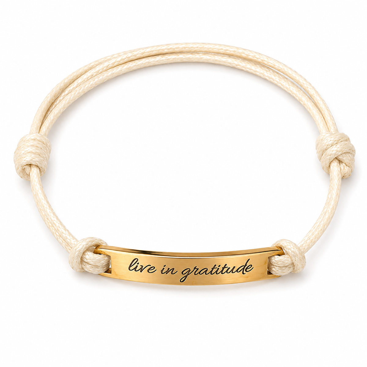 A double-strand cream-colored cord bracelet with a polished gold-plated ID bar engraved with the words 'live in gratitude' in black script, set against a plain white background.